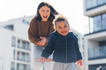 Laughing young caucasian mother chasing her little giggly biracial toddler son. Sunny day in the...