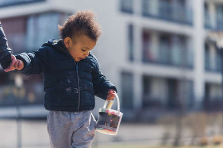 Brave little biracial baby boy holds a bucket of colorful chalks and holds his parents hand. Outdoor shot. High quality photoの写真素材