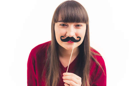 Attractive Caucasian young woman looks at the camera and holds fake moustache in front of her face movember white background copy space isolated studio shot . High quality photoの写真素材