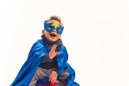 Studio shot of a little biracial boy toddler in a hero costume with a dark blue mask with a star and a cape laughing out loud over white background. High quality photoの写真素材