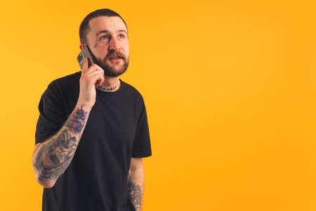 Medium studio shot of a caucasian bearded guy with tattoo sleeves wearing black t-shirt and talking via his smartphone over yellow background. High quality photoの写真素材