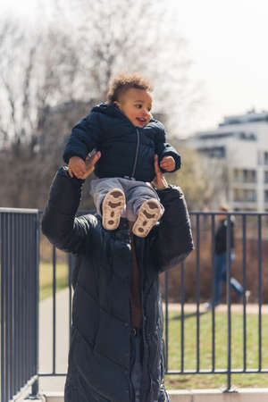 woman with a long black coat holding a cute curled little boy who is smiling upwards vertical outside medium shot. High quality photoの写真素材