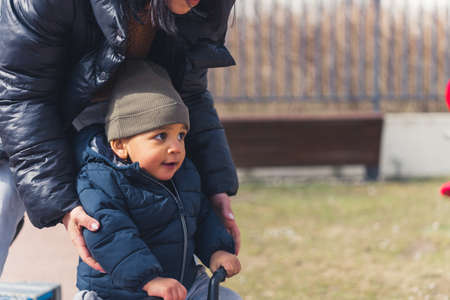 black-haired woman with a black coat touching a cute little boy with a grey hat who is sitting on a bike and looking ahead medium shot in the park outside. High quality photoの写真素材