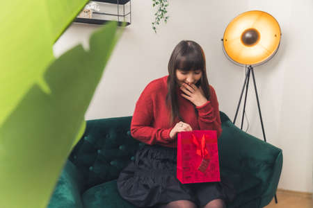 Excited young dark haired Caucasian woman opening a surprise gift copy space medium full shot home background . High quality photoの写真素材