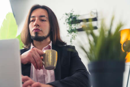 Medium closeup handsome young Caucasian man in a suit working at the office, holding a cup of coffee...