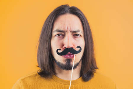 Movember concept - young frowned man holds fake paper moustache on a stick yellow background copy space isolated studio shot. High quality photoの写真素材
