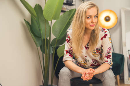 young blonde European girl in stylish clothes sitting on a stool and looking at the camera medium...
