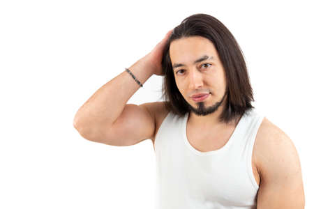 Caucasian bearded man in a white tank top brushing back his dark hair with one hand. Studio shot with white background. High quality photoの写真素材
