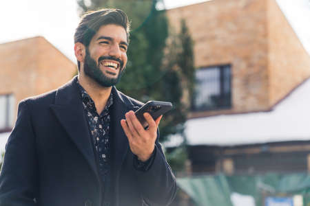 a young good-looking Latin American man in black clothes holding a phone and smiling widely medium closeup outdoor urban background . High quality photoの写真素材