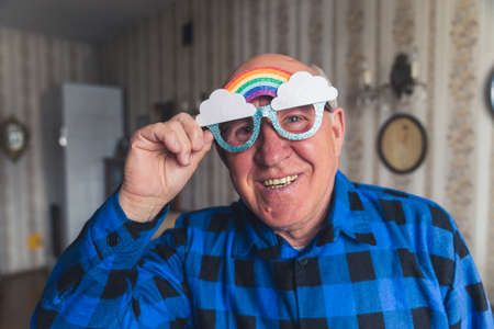Funny caucasian bald granddad looking at camera and smiling while wearing funky rainbow glasses. Vintage interior. High quality photoの写真素材