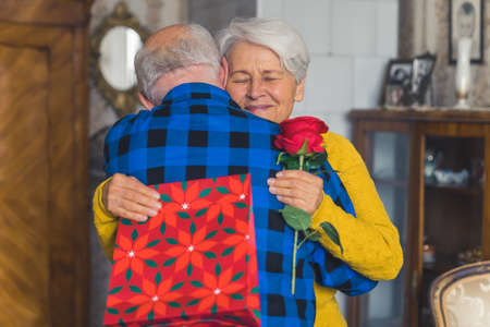 Two people embracing each other. Elderly caucasian couple celebrates birthday, valentines, or anniversary. Friendly grandmother is being hugged by her husband while holding presents. High quality photoの写真素材