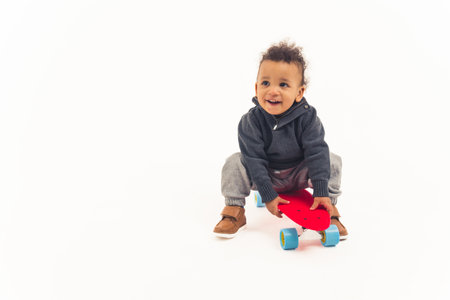 Biracial babys first skateboard ride. Curious toddler sitting on a red skateboard over white background. High quality photoの写真素材