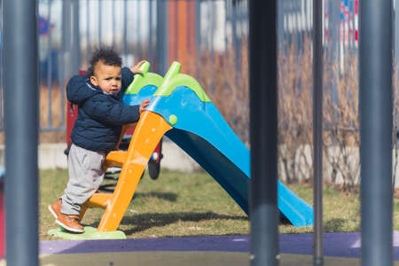 Curious toddler boy in warm clothes climbing on plastic playground slide. Springtime activities and happy childhood concept. High quality photoの写真素材