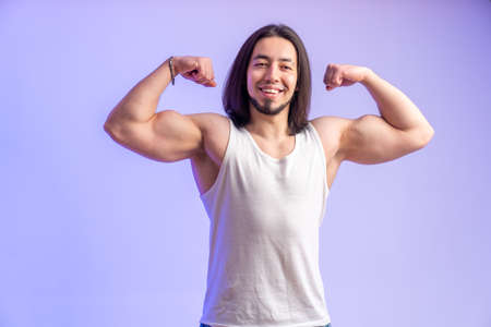 Young athletic man showing biceps and smiling and looking away studio shot medium shot isolated purple background indoors copy space . High quality photoの写真素材