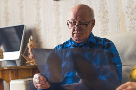 Concerned old wrinkled bald man in a blue flannel shirt and black glasses looking at his x-ray and worrying about his health. High quality photoの写真素材