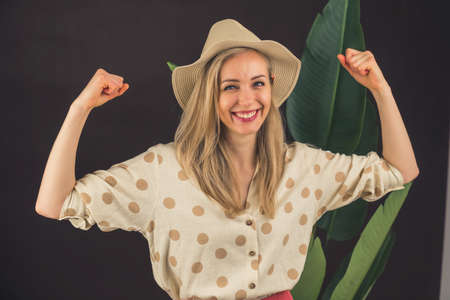 Smiling funny strong young blonde woman wearing white dotted shirt and hat spreading hands showing biceps muscles looking at camera. High quality photoの写真素材