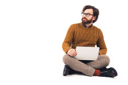 Bearded man wearing modern glasses sitting with crossed legs and holding laptop on the knees looking...
