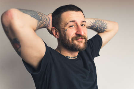 Happy bearded caucasian tattooed hipster man looking at camera and holding his hands in the back of his head. Medium studio shot over gray background. High quality photoの写真素材