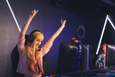 Victory. Excited satisfied long-haired female gamer influencer wins her next online match in a tournament. Happiness shown by throwing hands in the air. Internet cafe. High quality photoの写真素材