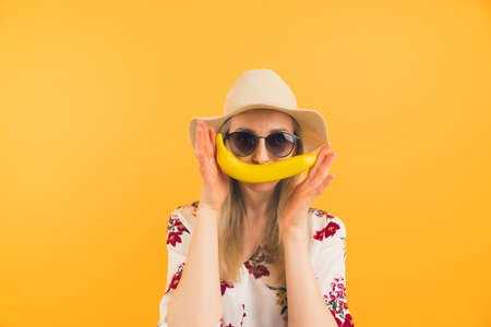 young blonde girl with a hat and sunglasses holding a banana as a mustache medium closeup studio shot orange background healthy lifestyle concept. High quality photoの写真素材