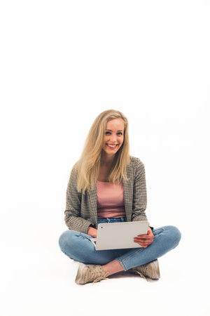 nice European blonde girl sitting with crossed legs on the floor laughing and holding a laptop full shot studio shot white background copy space vertical . High quality photoの写真素材
