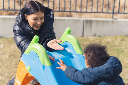 Mother - son relationship. Outdoor shot of a young caucasian black-haired mum encouraging her biracial toddler boy to catch her hands and climb the slide. High quality photoの写真素材