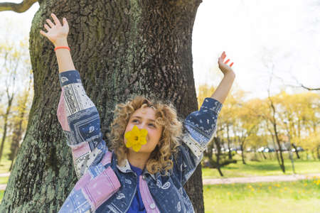 Attractive blonde lady with curly hair and with yellow flower in her mouth raising her hands in the...