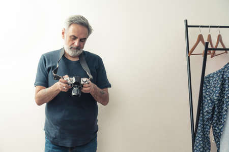 Medium studio shot. Elderly casually dressed grey-haired man holding a professional camera in his hands, standing near a clothes rack. High quality photoの写真素材