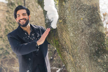 Stylish Handsome Latin Man in black coat and suit playing hide and seek behind the tree in a park...