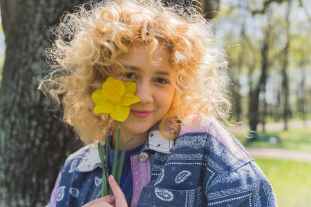 Caucasian blonde curly-haired cute girl in a park holding a yellow flower near her face. Outdoor...
