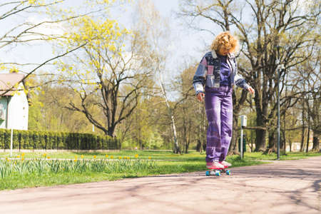 Concentrated attentive woman in her 20s with curly hair carefully riding a skateboard in a park...