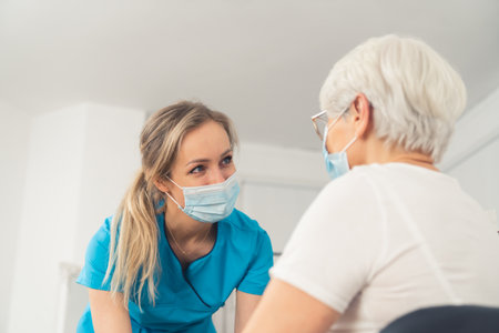 Young caucasian female nurse helping and looking at an elderly woman, both are wearing masks. Helthcare concept. Inner shot. High quality photoの写真素材