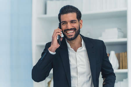 Indoor portrait of a laughing handsome dark-haired bearded Cuban businessman in a suit talking to...