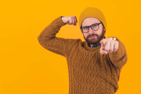 Funny serious hipster caucasian man with a beard, glasses, and a beanie standing over yellow background pointing at camera and tightening his muscles. High quality photoの写真素材