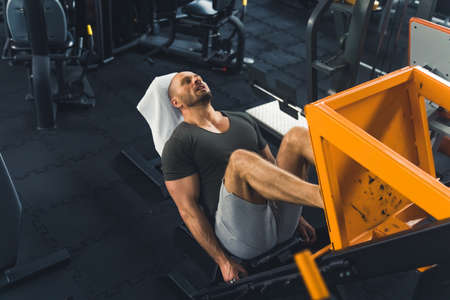 Extremly tired bald well-built man lying on a bench exercising on a leg press. Indoor shot at a well-equipped gym. High quality photoの写真素材