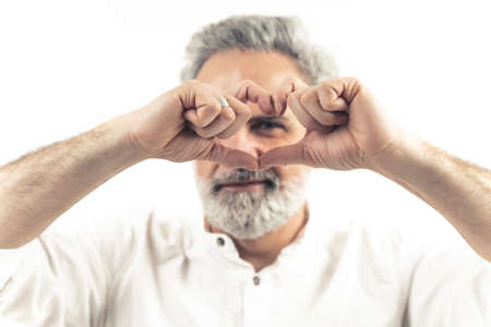Studio shot on white background of an european attractive man in his 60s making a heart sign with his fingers over his face. Focus on the foreground. High quality photoの写真素材