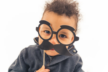 medium close up shot over white background of an interracial cute toddler baby boy holding black fake paper moustache and glasses. happy childhood concept. High quality photoの写真素材