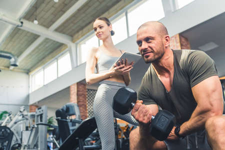 Female attractive slim personal trainer coordinating a weight training of a middle-aged bald strong guy lifting a 10 kilograms dumbbell. High quality photoの写真素材