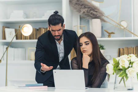 Brunette pretty elegant caucasian businesswoman sitting on a chair in front of a laptop. Unhappy bearded cuban man poiting at the screen looking dissatisfied. High quality photoの写真素材
