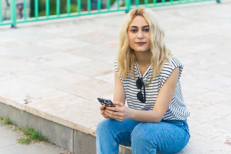 blonde Caucasian girl reading good news into the phone and smiling medium full shot outdoor. High...