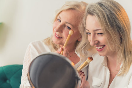 Close-up indoor shot of two blonde attractive millenial cheerful women having fun while doing make-up in front of a portable round mirror. High quality photoの写真素材