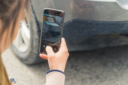 Outdoor shot of an unrecongnizable caucasian woman taking a photo on a road of her cars front fender with a modern smartphone. High quality photoの写真素材