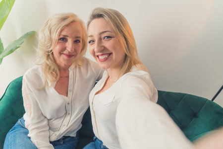 Indoor shot of two related caucasian women in their 40s wearing white elegant shirts takinga selfie, hugging each other and smiling. High quality photoの写真素材