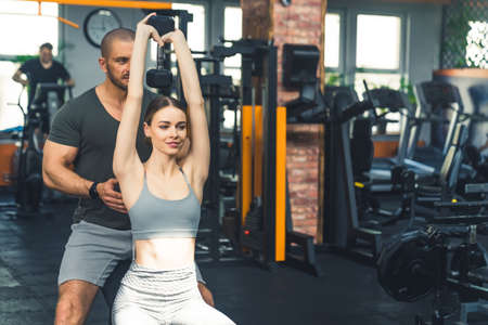 Beautiful dark-haired slim woman in her 20s sitting on a bench and lifting a heavy dumbbell with her...