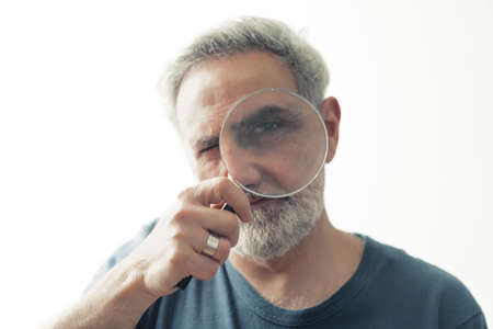 grey haired bearded man holding magnifier in front of his face white background closeup studio shot isolated . High quality photoの写真素材