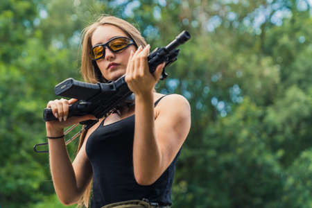 Outdoor shooting range concept. Young beautiful long-haired caucasian girl in a black tank top looking up close at a black rifle. Blurred trees in the background. Medium shot. High quality photoの写真素材