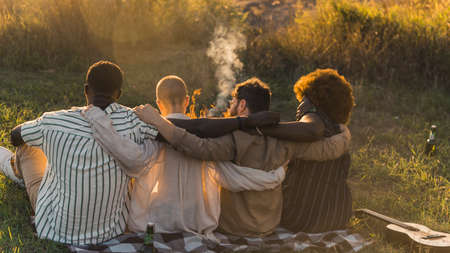 Rear view of multicultural good old friends hugging while sitting on a blanket on the grass, having a weekend picnic in nature near the river, talking and drinking beer. Sunset time. High quality photoの写真素材