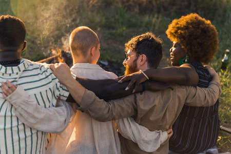 Rear view of multicultural good old friends hugging and having a weekend picnic in nature near the...