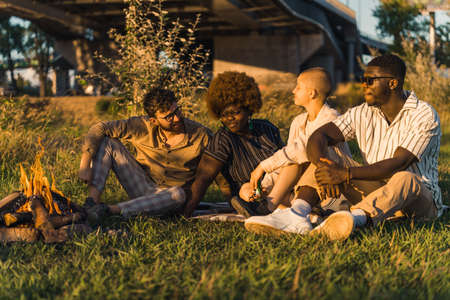 Multi-ethnic group of good friends enjoying their summer picnic in nature while sitting on the grass near the fire, drinking beer, talking and watching the sunset. High quality photoの写真素材