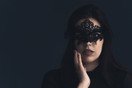 White woman with dark long hair and a black lace mask looking into camera and touching her cheek. Dark background studio shot. High quality photoの写真素材